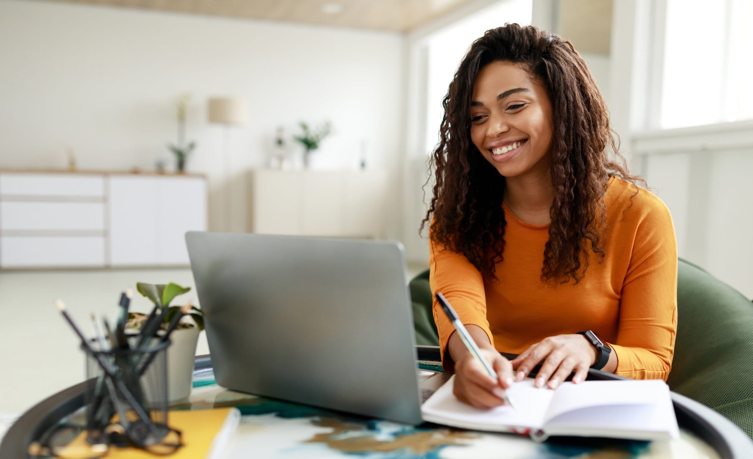 Black woman sitting at desk, using pc writing in notebook QLS logo for an accreditation online course